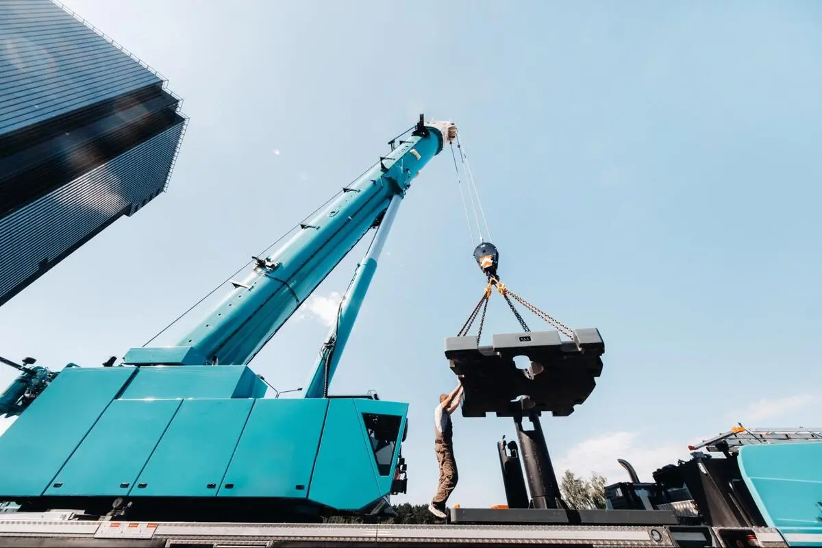 Teal mobile crane lifting a load beside city high-rise buildings